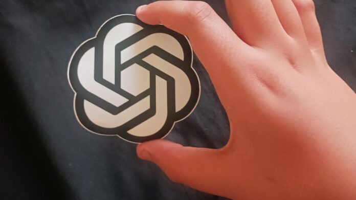 A hand holding a sticker with the OpenAI logo against a dark fabric background